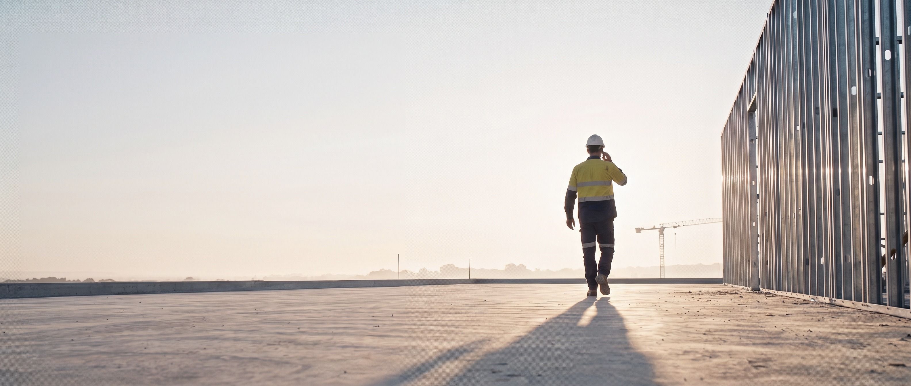 A foreman walks away across the slab toward the rising sun with a phone to his ear, his shadow long behind him.